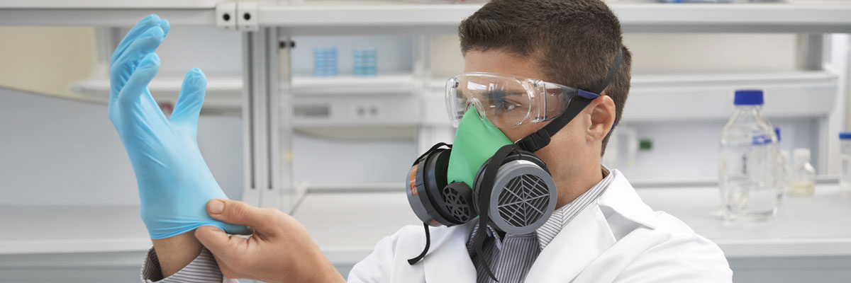 A student in a lab coat, safety glasses and respirator dons nitrile gloves in a lab.