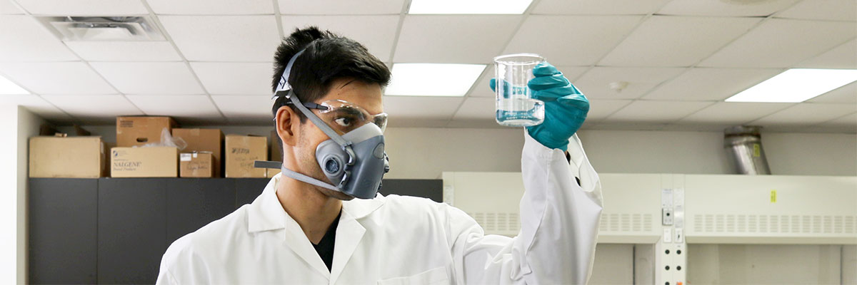 A man in a lab coat, safety glasses, nitrile gloves and a respirator examines a beaker full of a clear fluid.