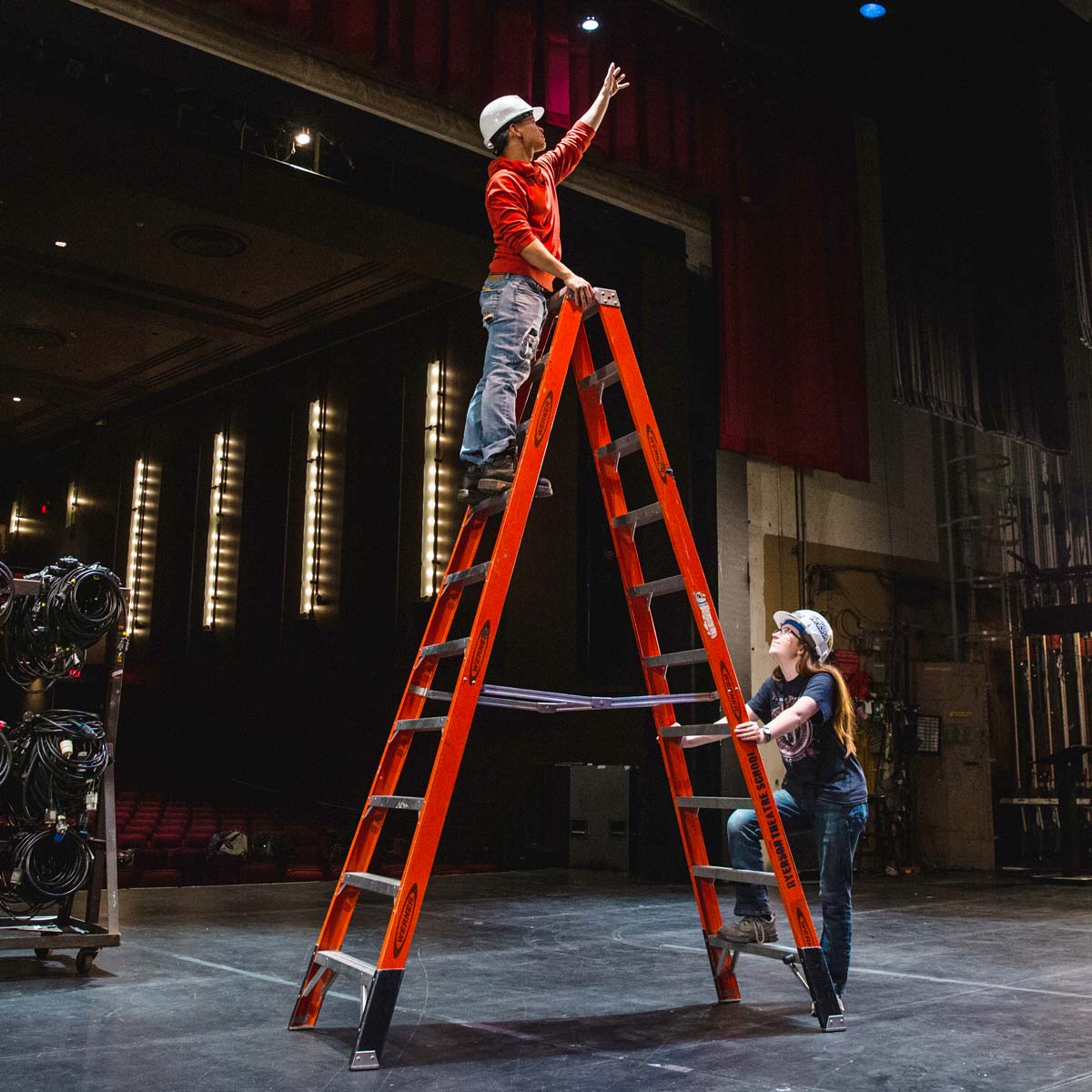 A student is using a ladder to hang lights in the theatre and is maintaining three points of contact with the ladder.