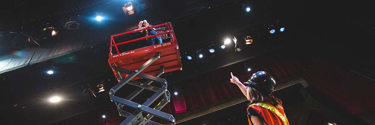 A student is being raised up on a scissor lift to access lights that are high above the stage in the Kerr Hall theatre.