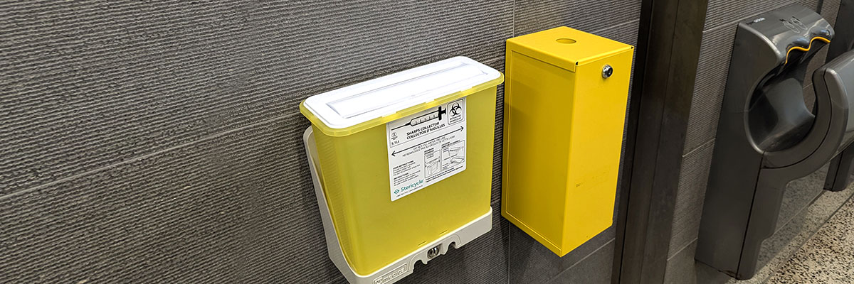A large sharps disposal container is mounted on the wall next to a standard-size container in a Kerr Hall washroom.