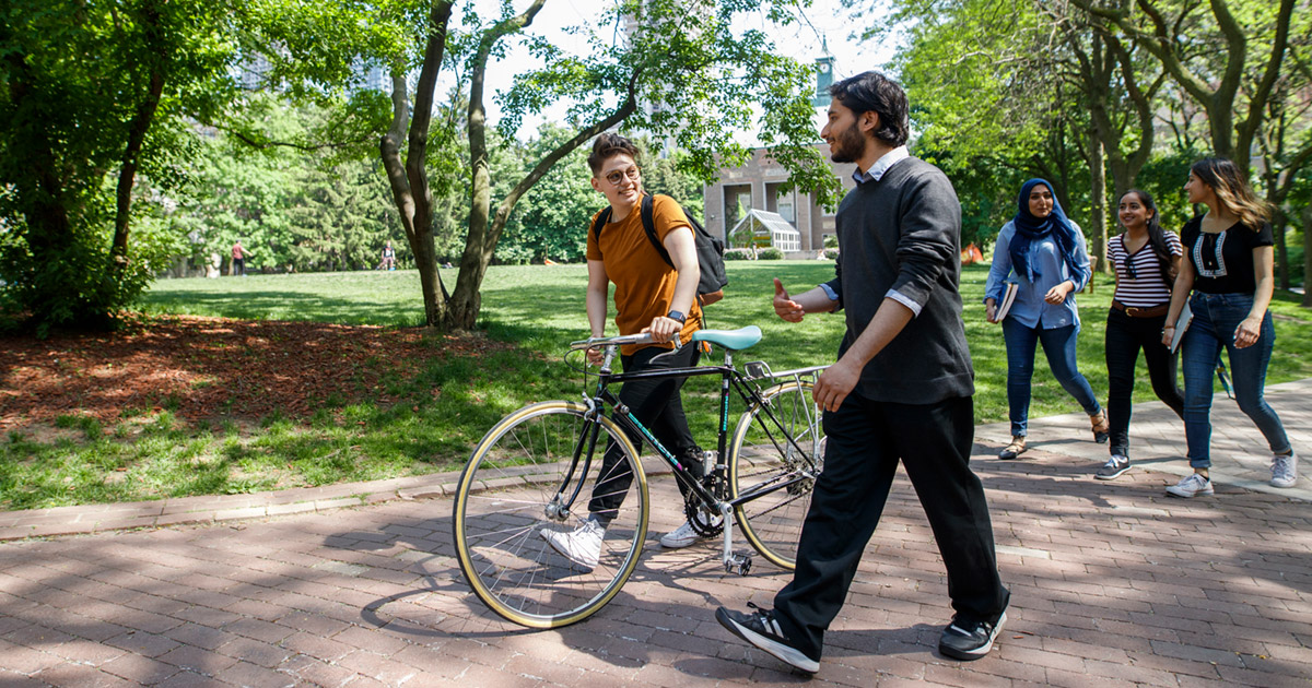 Five students walking through the lush green Quad, one of them with a bicycle.