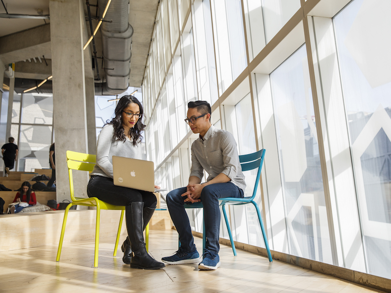 Two individuals sit on chairs side by side and look at a laptop together