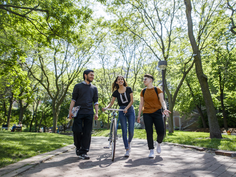 Three students are walking and engaging in a conversation with each other at the quad. One of the students is walking with her bike while another holds a laptop.