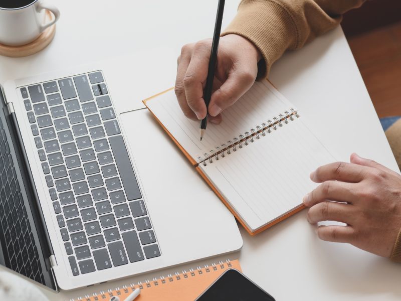 A close-up of a person writing in a notebook with their laptop.