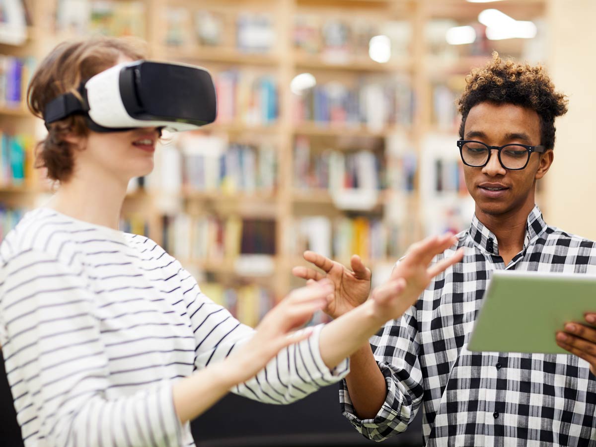 A person gestures while using a white and black VR headset against a backdrop of books as another person stands next to them, appearing to give directions while holding a green book. 