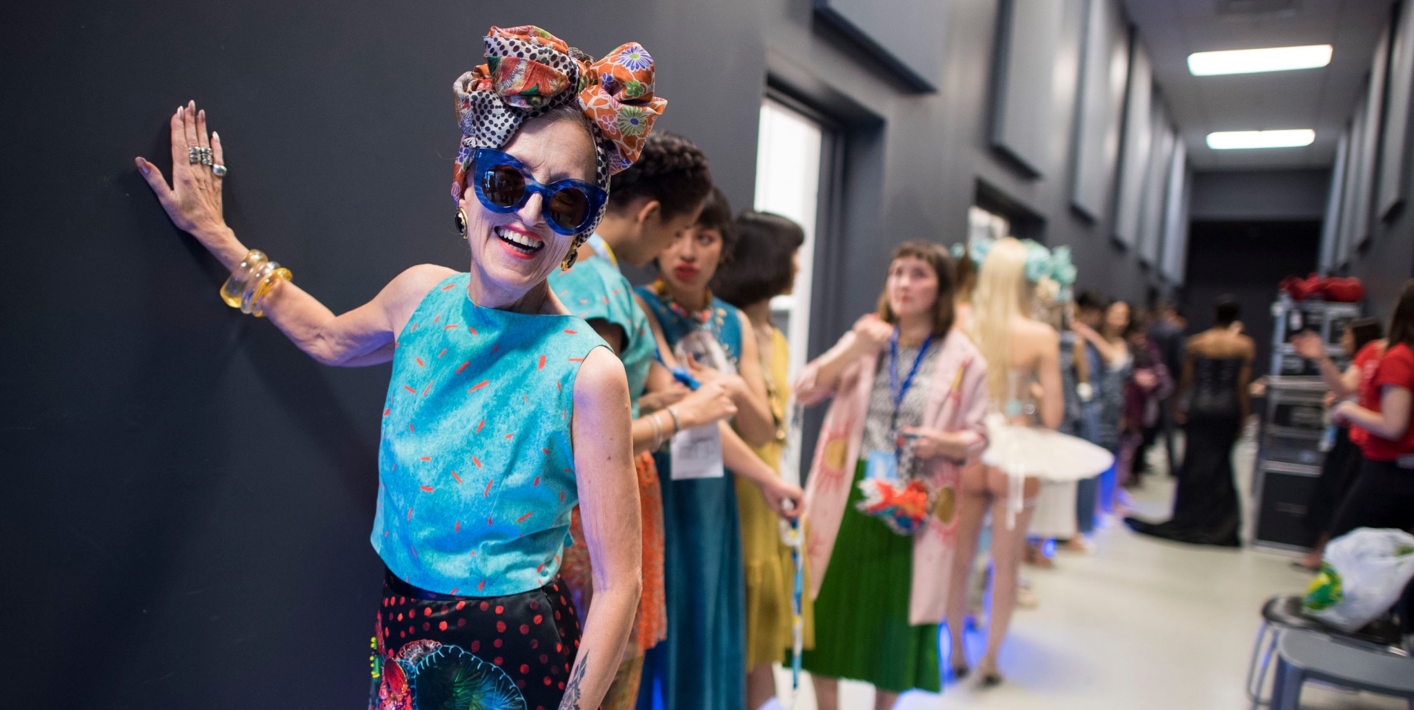Group of models lined up backstage, with one woman standing happily in colourful clothing and headscarf