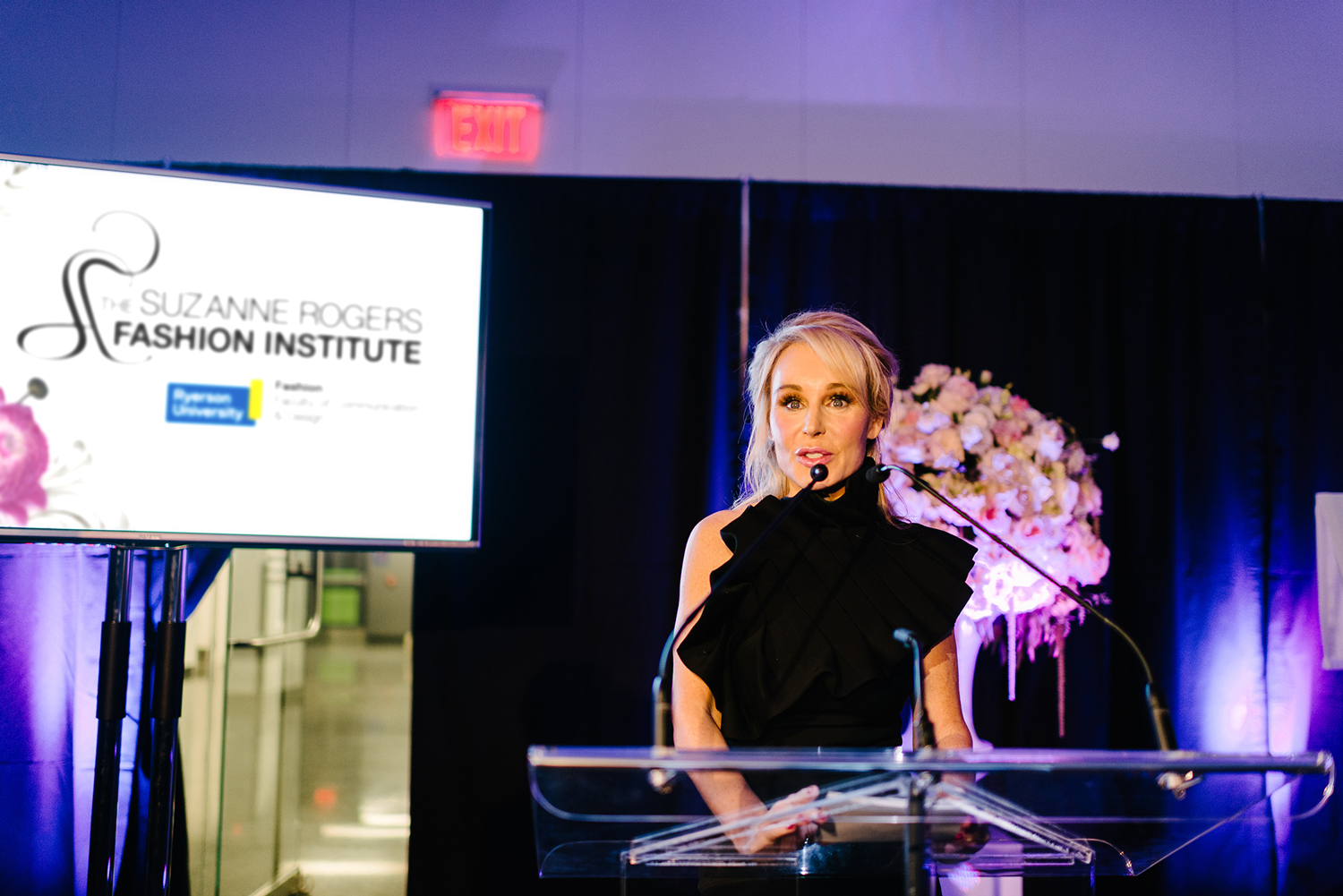 Suzanne Rogers on stage announcing the inauguration of The Suzanne Rogers Fashion Institute wearing a black dress.