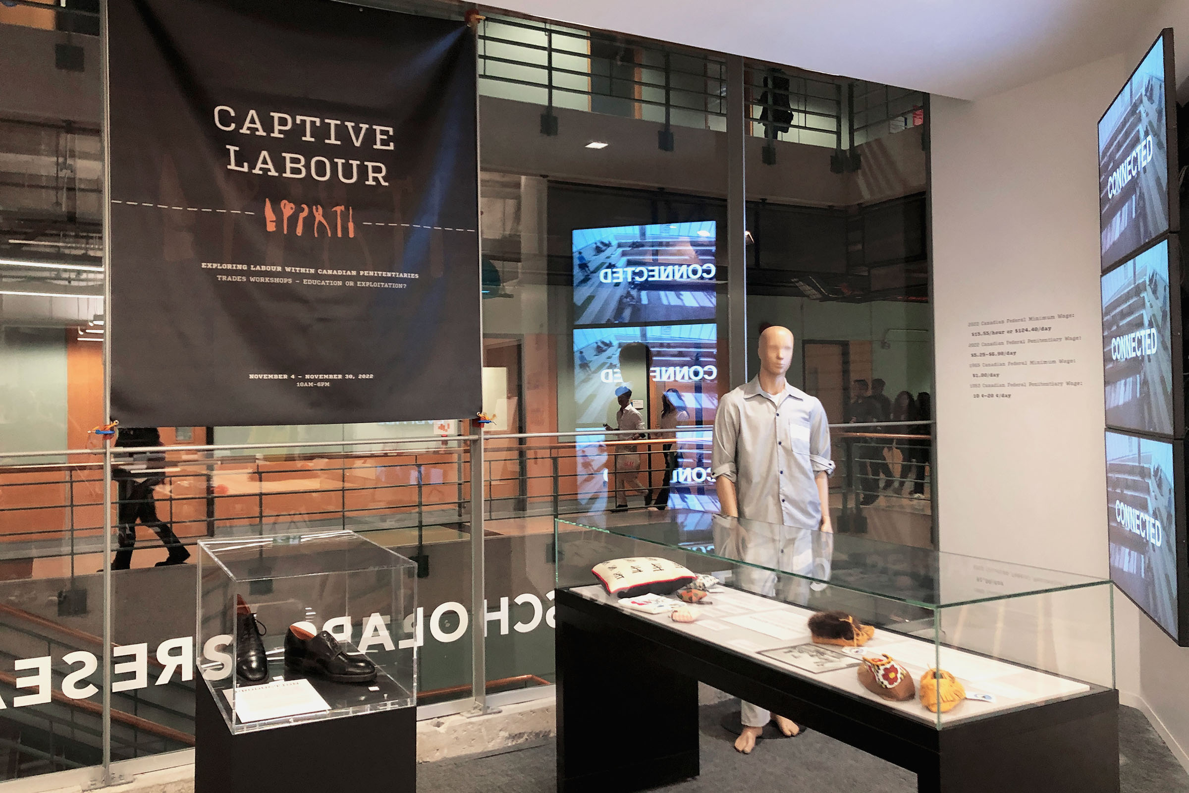 Landscape photo of Captive Labour exhibition, showcasing two glass cases and a mannequin wearing a prison uniform behind them. Three screens on the right side of the frame display facts about prison labour 