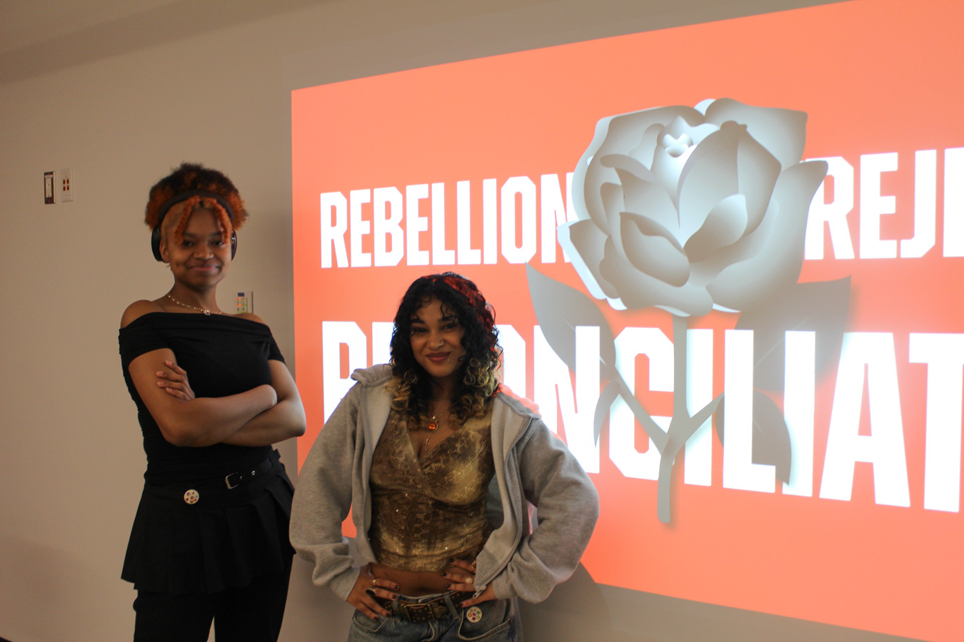 Two students stand in front of a projected photo on a white wall that reads ‘Rebellion Rejection Reconciliation’ in blocky white text on a red background there is a rose in the center of the three words 