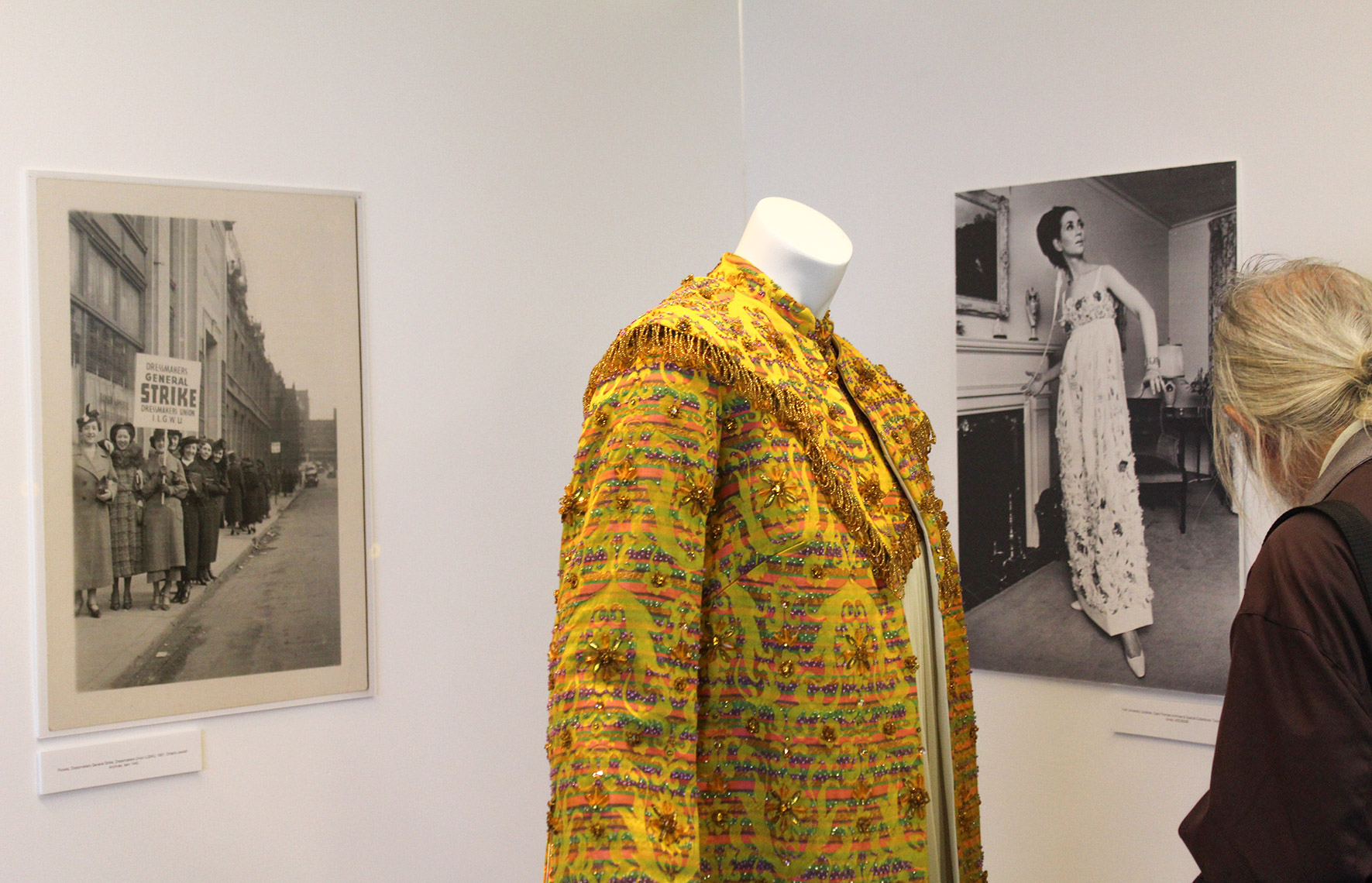 Close up of Ruth Dukas garment in between the ‘v’ of two white panels, each panel has a grayscale photo mounted on it. There is someone looking at the garment on the right side of the image. 