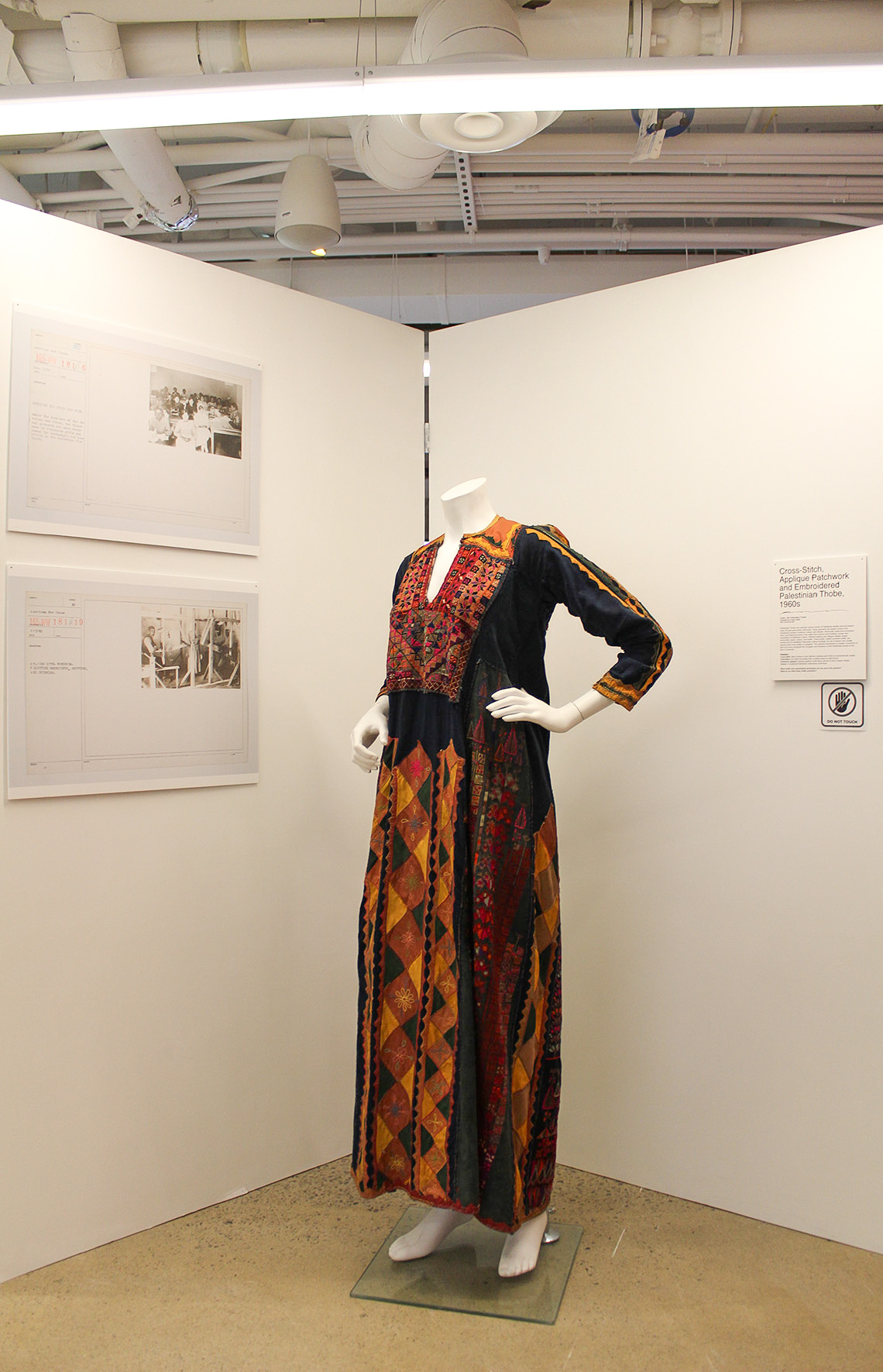Palestinian thobe sits at the centre of two white panels that form a ‘v’ shape. There are two plaques on the left panel, and one plaque on the right. 