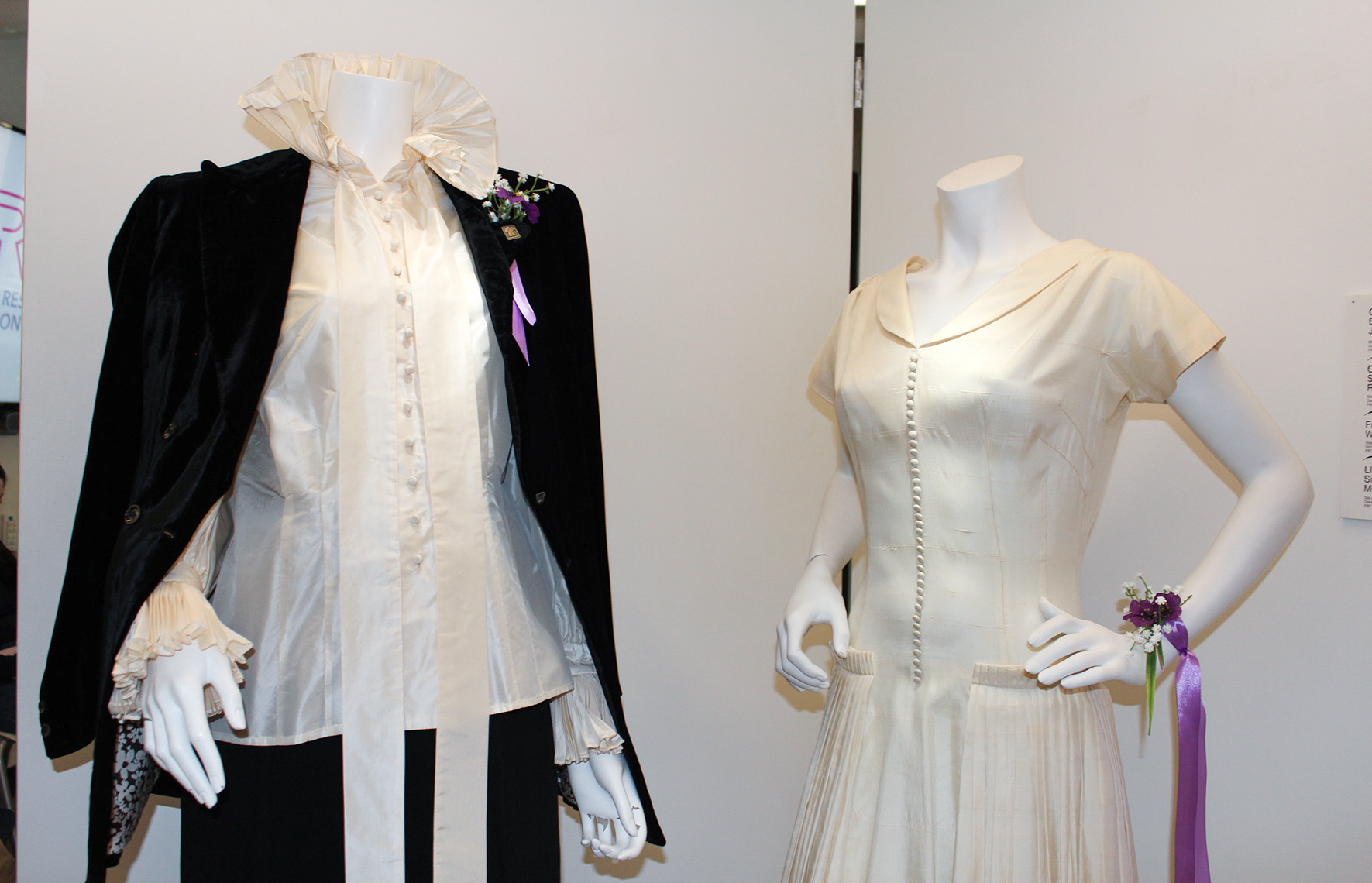 Close up of two mannequins, one dressed in a suit and the other in a dress. They have matching purple corsages and boutonnières. 