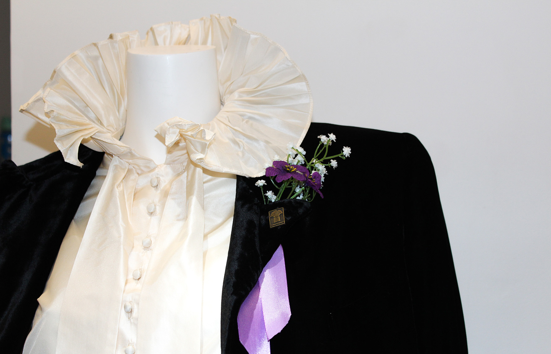 Close up of mannequin wearing a ruffled blouse and suit jacket. A purple boutonnière is pinned to the left lapel of the suit jacket. 