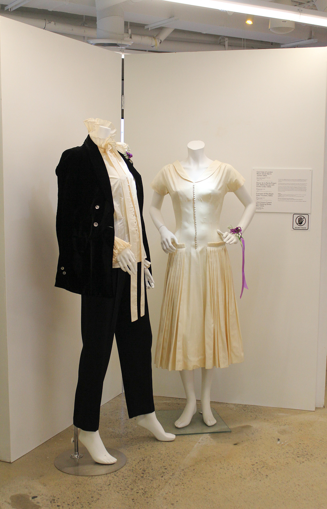 Two mannequins, one wearing a suit and one wearing a dress stand in between two white panels that form a ‘v’. 