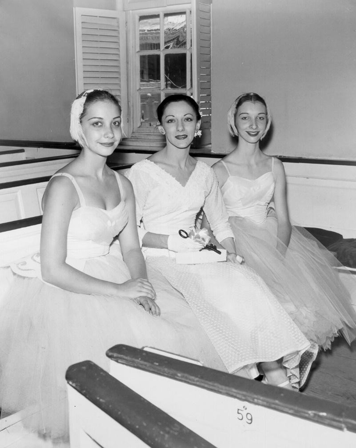 Celia Franca sitting in between two ballet dancers on a US tour, 1956. All three women are wearing light colours. The photo is in black and white. 