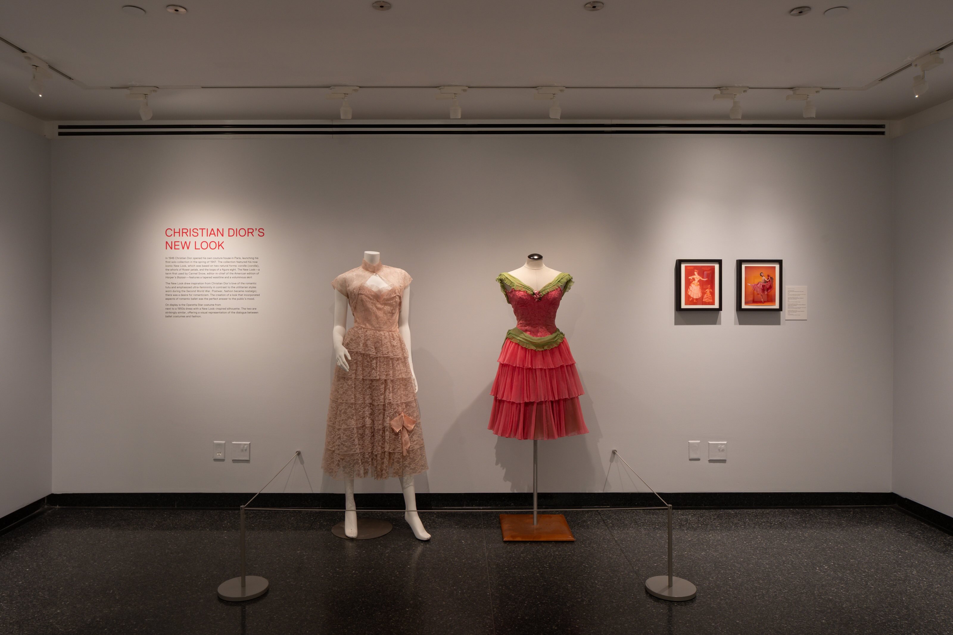 A wall with exhibition text to the left, two dressed dress forms in the centre: the left one wears a pale pink 1950s dress with a matching bolero, the right wears one of Celia Franca’s ballet costumes. Two framed photos are on the right. 