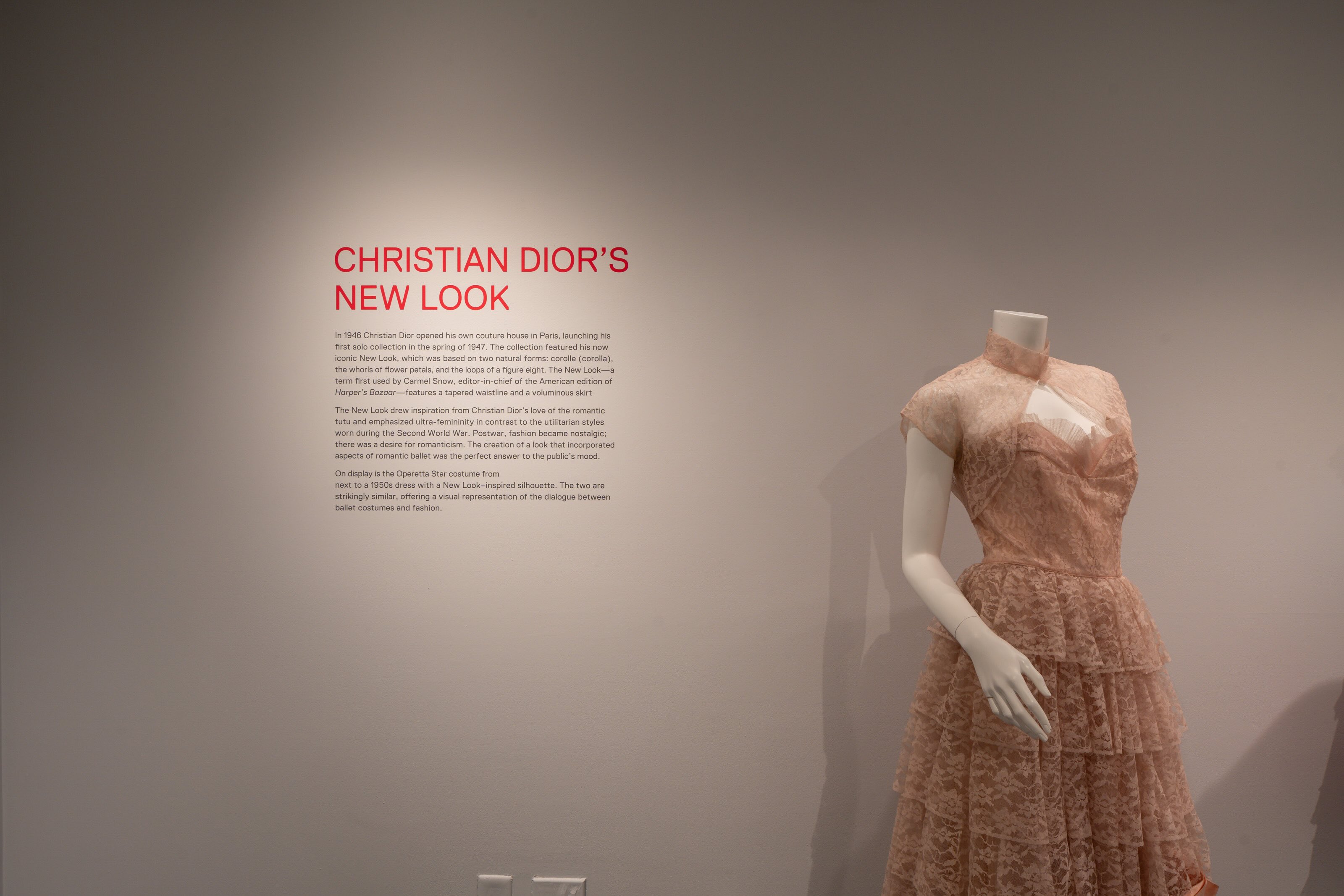 A wall with exhibition text and a white mannequin dressed in a pale pink dress with a bolero to the right. Heading reads “Christian Dior’s New Look” in bright pink text. 