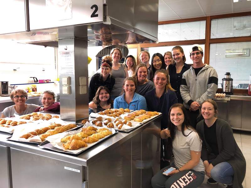 Food and nutrition students smiling together in kitchen, in front of a variety of baked goods.