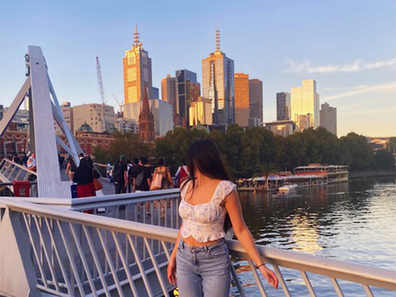 Laura Pietroiusti looks away from the camera while leaning on a boardwalk railing with a city skyline behind her