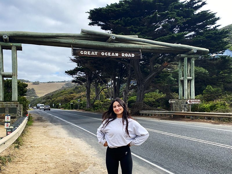 Laura Pietroiusti poses for the camera next to a road with a road sign saying "Great Ocean Road" behind her