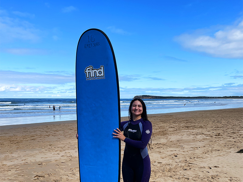 Laura Pietroiusti dressed in a wet suit poses beside a surf board on a beach in Australia.