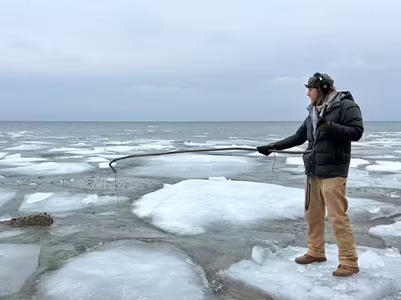 A man wearing headphones holds a boom mic out over a lake as he records sounds of ice floes.