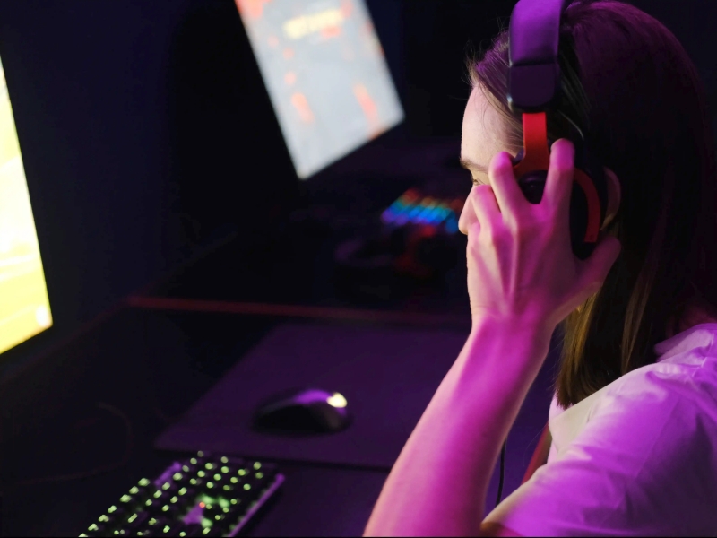 A woman holds her headset while bathed in the glow of a computer monitor
