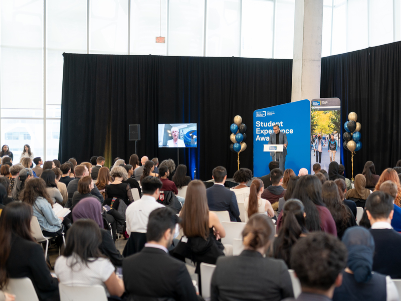 A presenter at the podium speaks to an audience a large audience in the SLC student learning centre