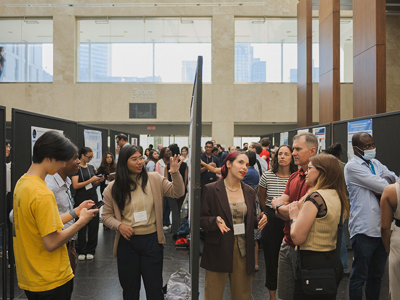 Large diverse group of people standing in a hall with boards depicting various student research projects. They discuss in small groups the research.