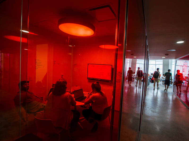 Three people meet in a red-lit glass conference room as others walk through a bright, modern corridor in the Student Learning Centre.