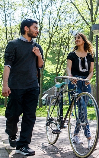 Three students walk together on a sunlit path in the Quad. One student is walking with a bike while another carries a laptop.