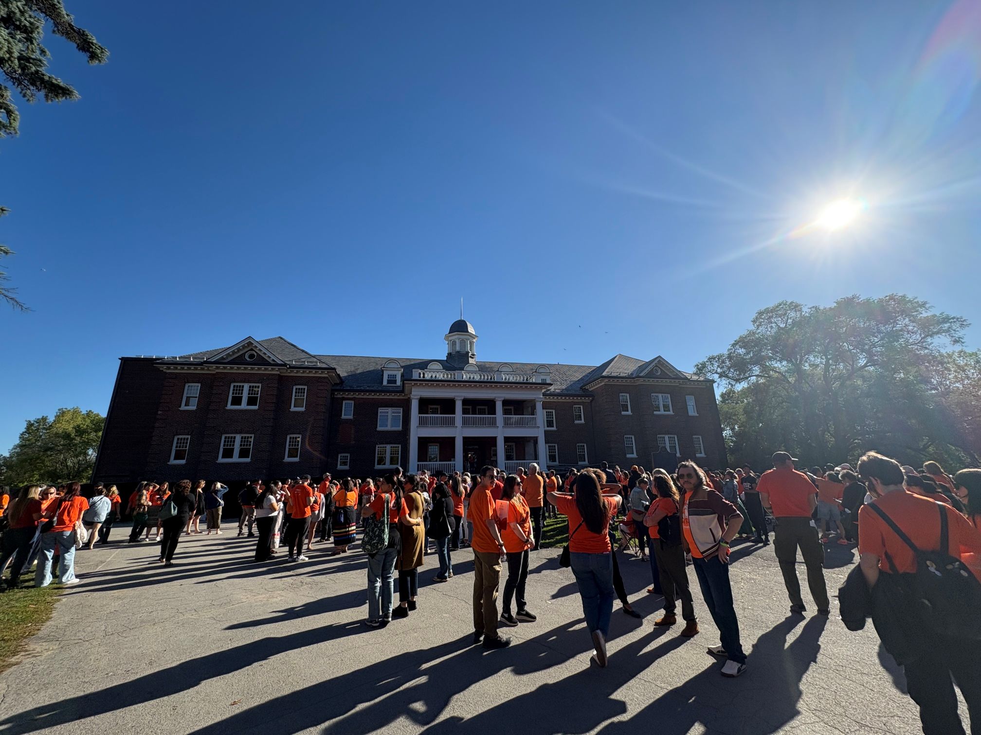 A group of people in orange shirts gather outside the former Mohawk Institute Residential School while the sun shines overhead.