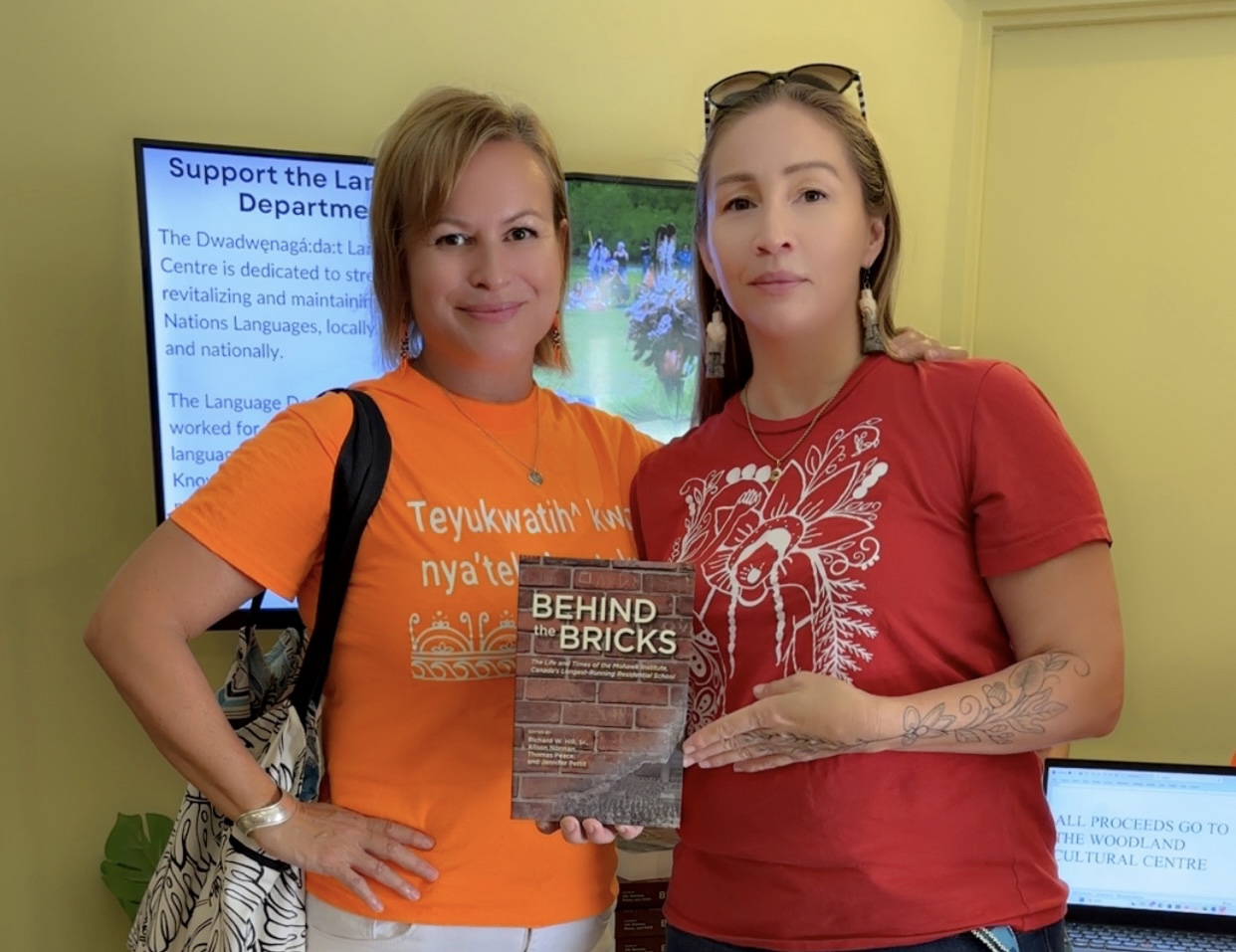 Dr. Sandra Juutilainen and Teri Lyn Morrow, both wearing orange shirts associated with reconciliation toward Indigenous people, hold a copy of the book Behind the Bricks.