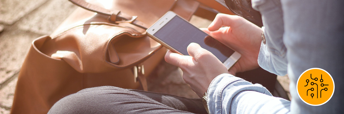 A person looking at their cellphone sitting on the ground outside with their brown leather bag beside them.