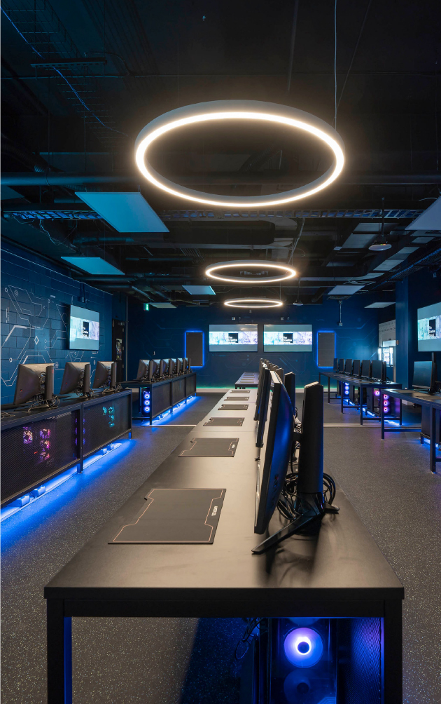 Gaming computers sitting on desks in a row under a futuristic, circular lamp.