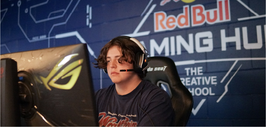 Student with gaming headphones playing video game on a computer front of the Red Bull Gaming Hub sign.