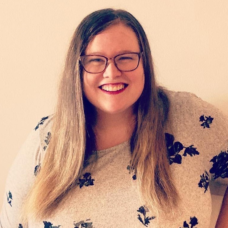 Meghan smiles at the camera. She has long hair and glasses. Her right hand is on her hip.