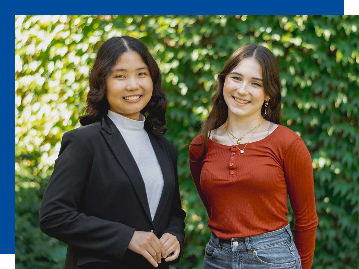 Two young women smile while standing in front of a wall covered in green vines.