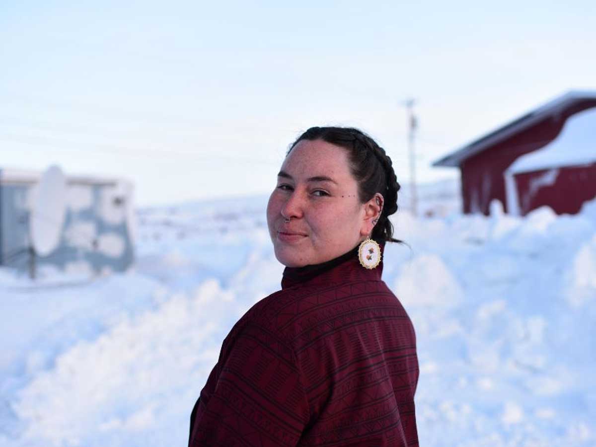 Paige Kimiksana Kreps looks back at the camera framed by two outbuildings in landscape of deep snow.
