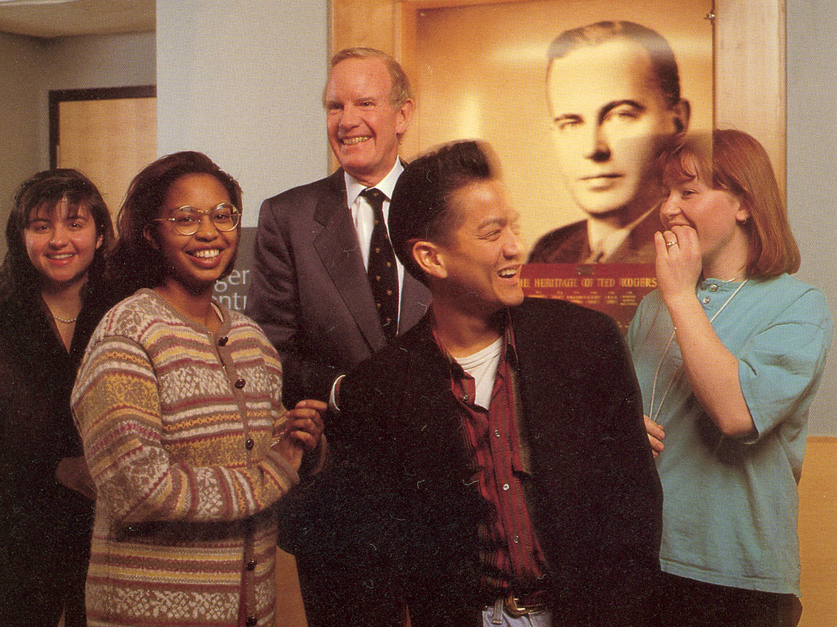 Ted Rogers Jr. smiles and stands among members of the Toronto Metropolitan Community in front of a photo of his father, Ted Sr.