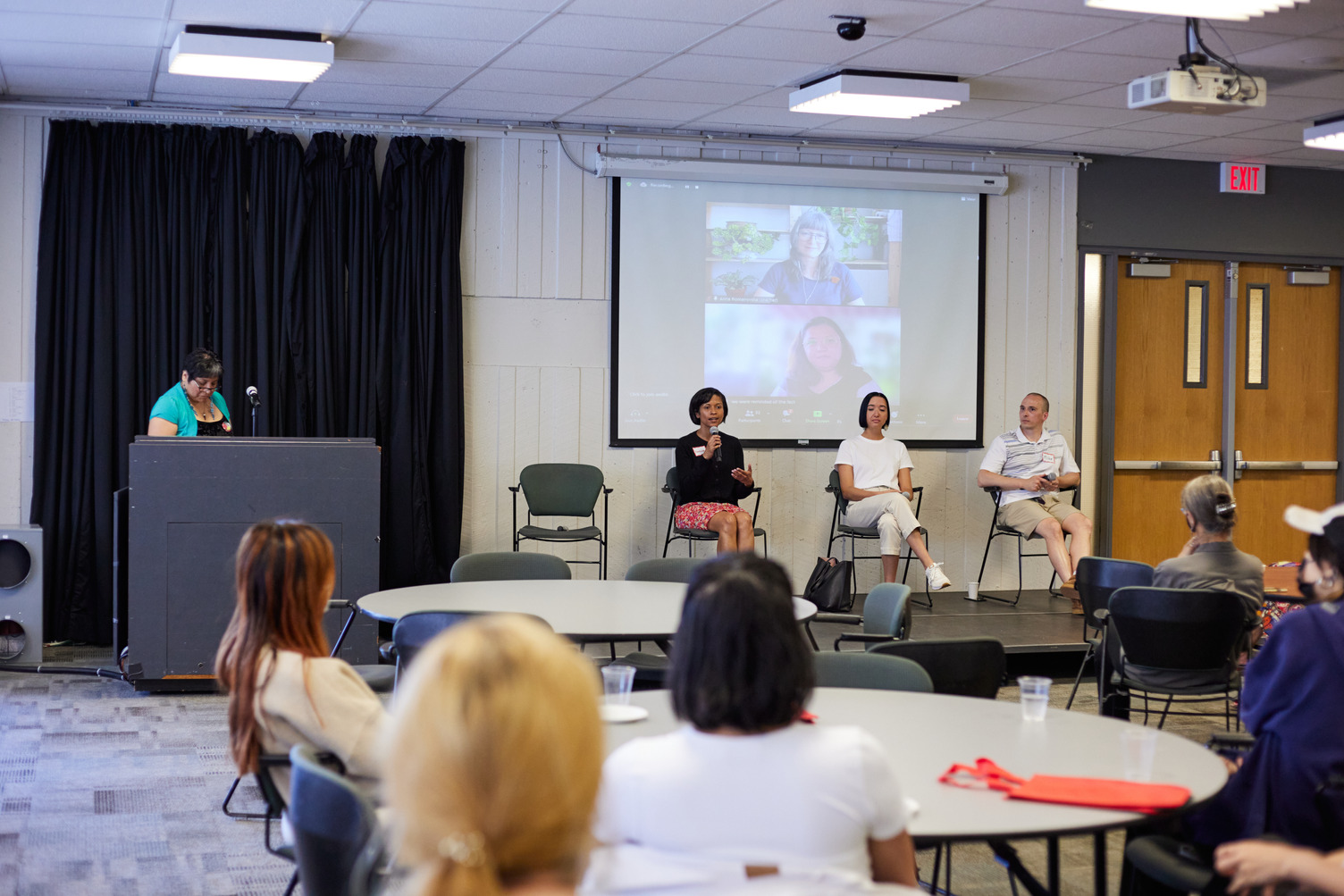 A hybrid panel discussion with in-person speakers and virtual participants projected on a screen.