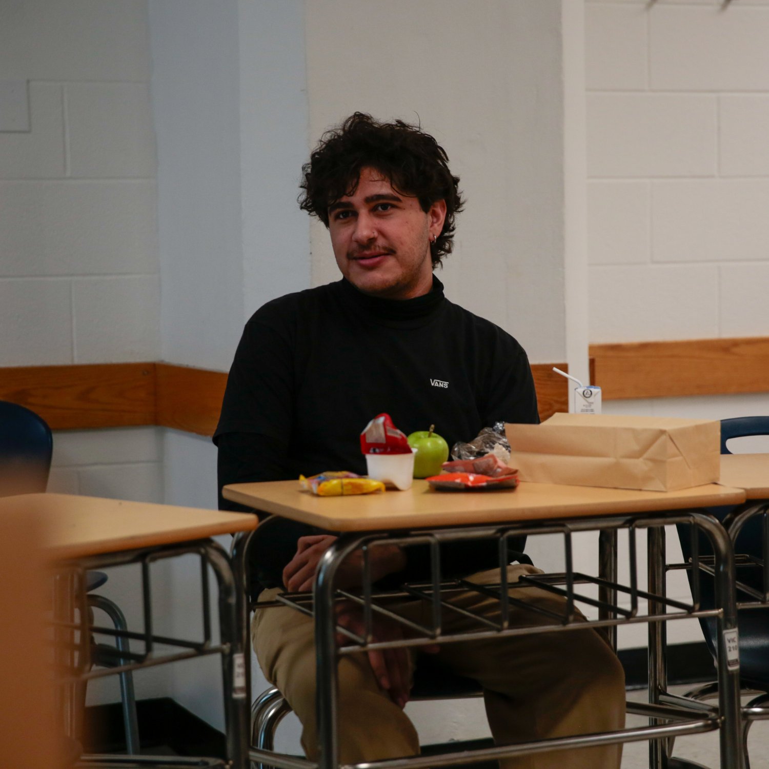 A student sitting at a desk with a brown bag lunch and snacks in a classroom setting.