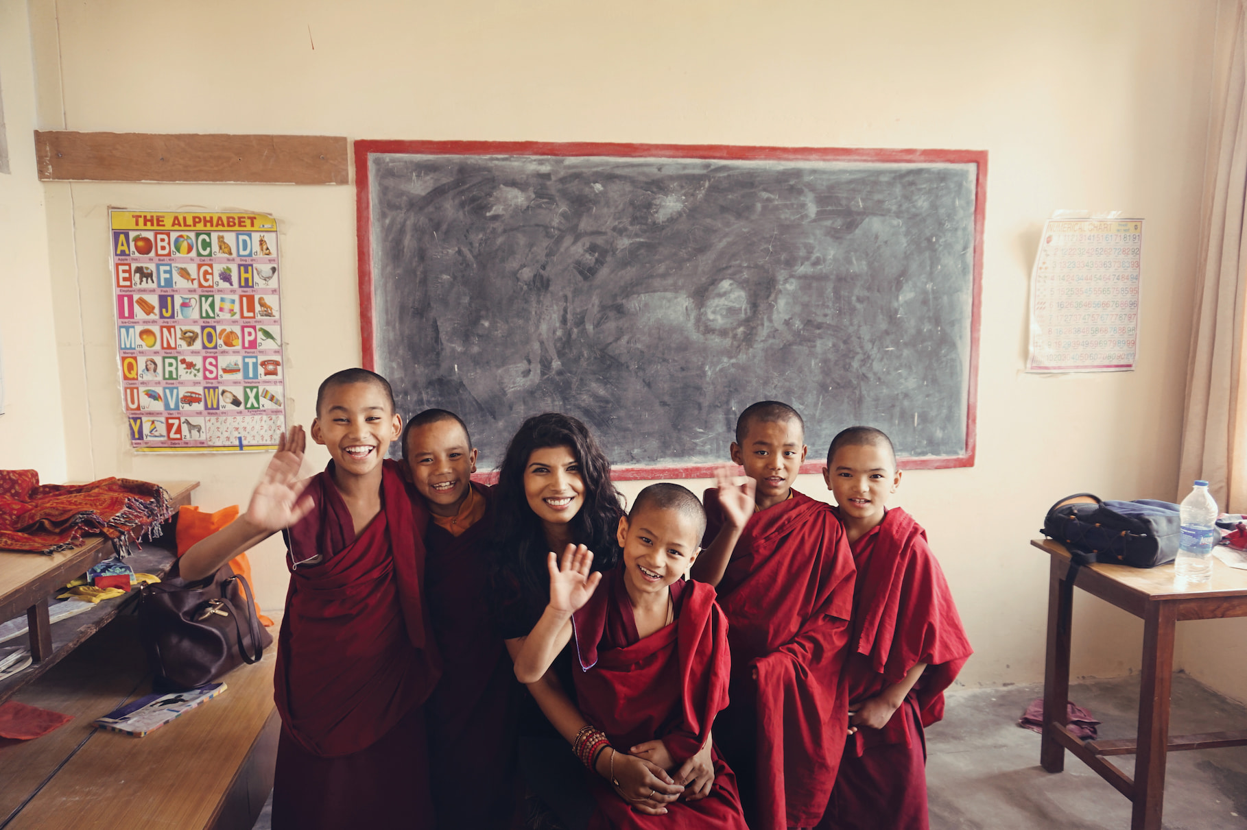 Smiling student sitting with a group of young monks in red robes inside a classroom, posing together in front of a chalkboard and waving at the camera.
