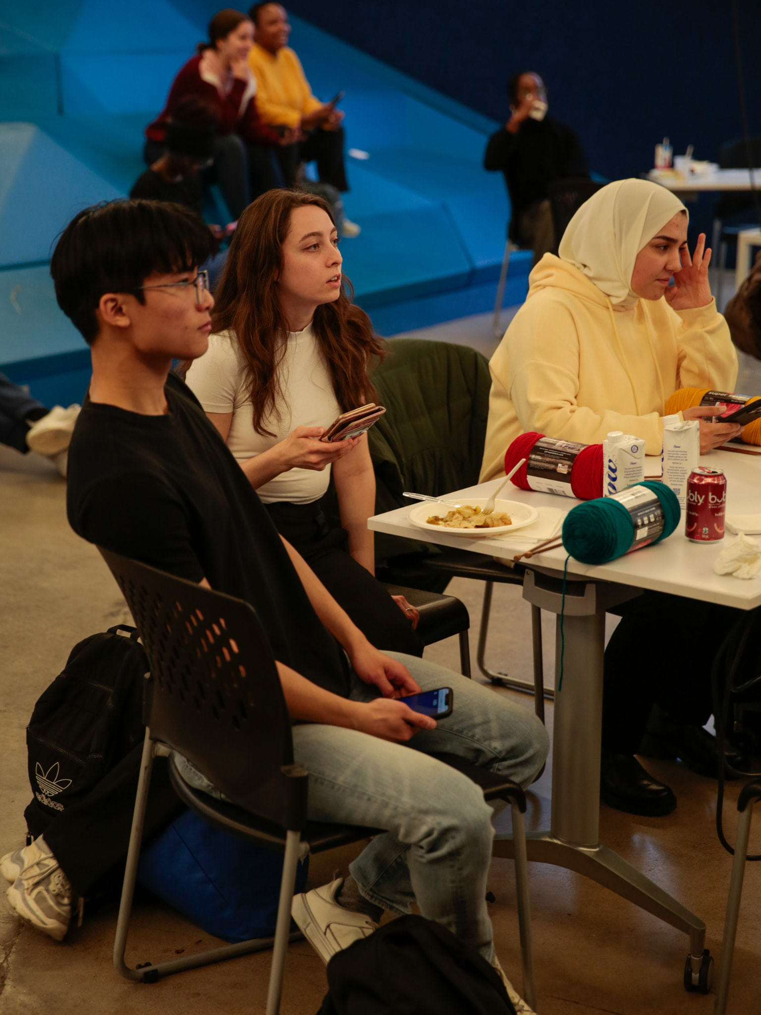 Students seated indoors with food, drinks, and yarn, engaging in a casual campus activity.