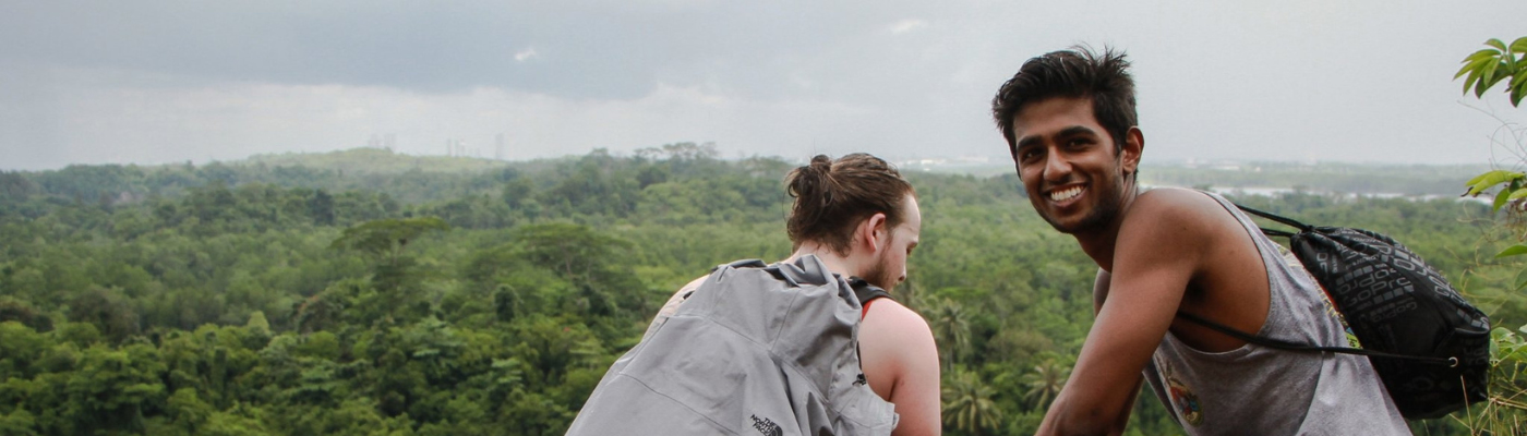 Two young men at the edge of a cliff overlooking a lush green forest and a lake. One is crouched looking down, wearing a gray jacket, while the other smiles at the camera, wearing a tank top and backpack.