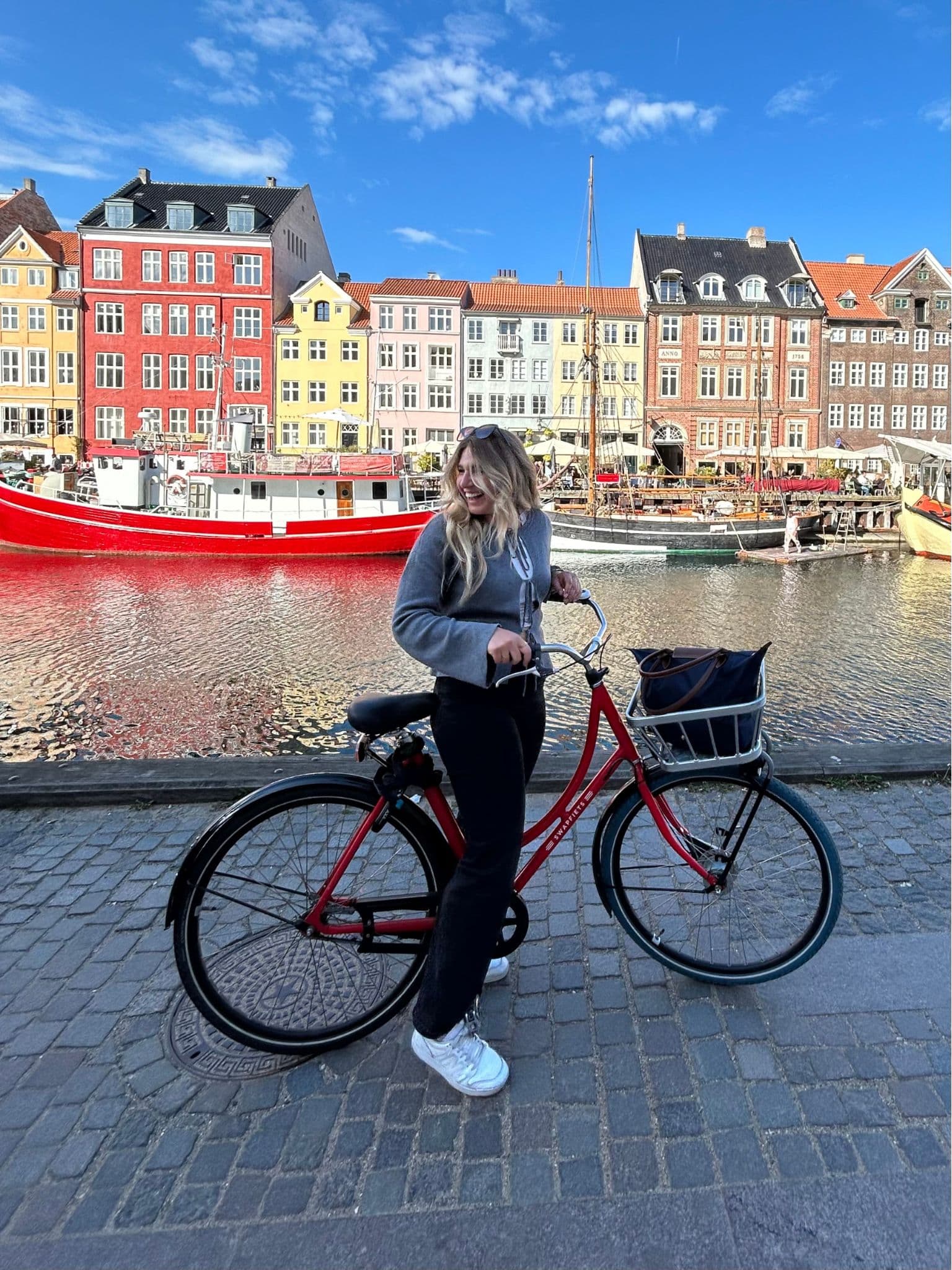 A woman in a gray sweater rides a red bicycle on a cobblestone street by a canal, with colorful buildings and boats in the background under a blue sky.