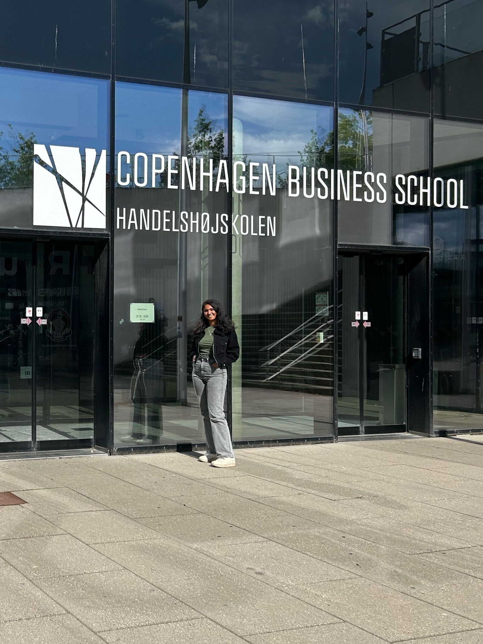 A person stands in front of the glass entrance to Copenhagen Business School, with the schools name and logo displayed above the doors. Its a sunny day and the pavement outside is well lit.