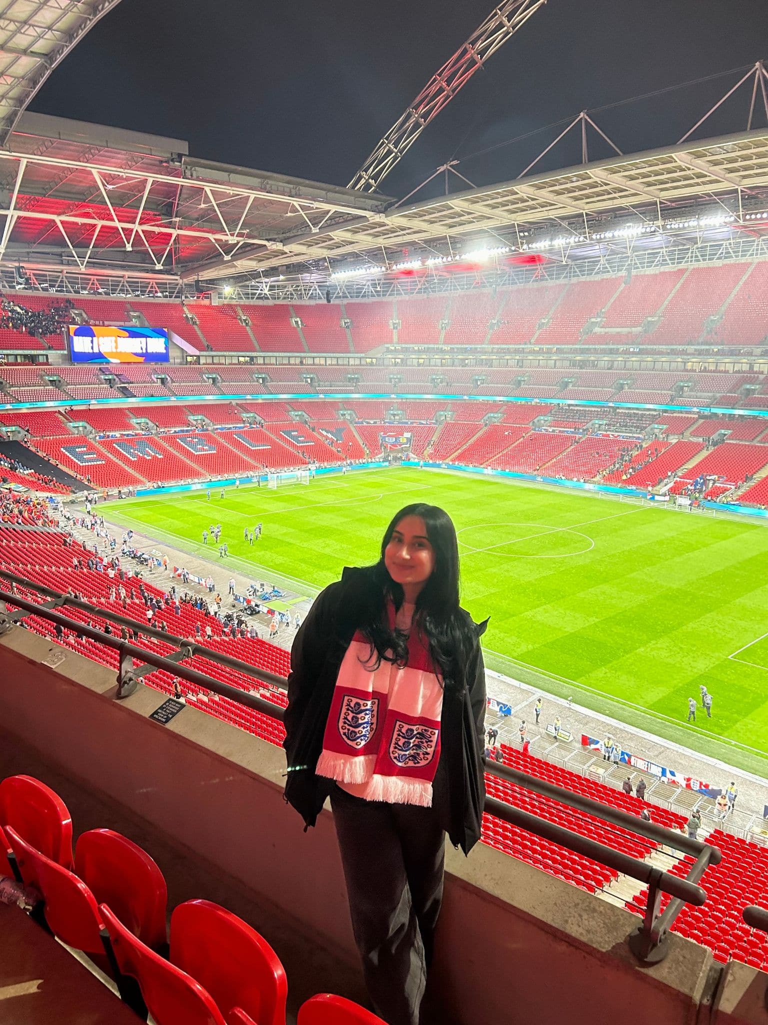A woman wearing a white scarf with England football logos stands smiling in a stadium with bright lights and mostly empty red seats. The pitch and a few people are visible in the background.