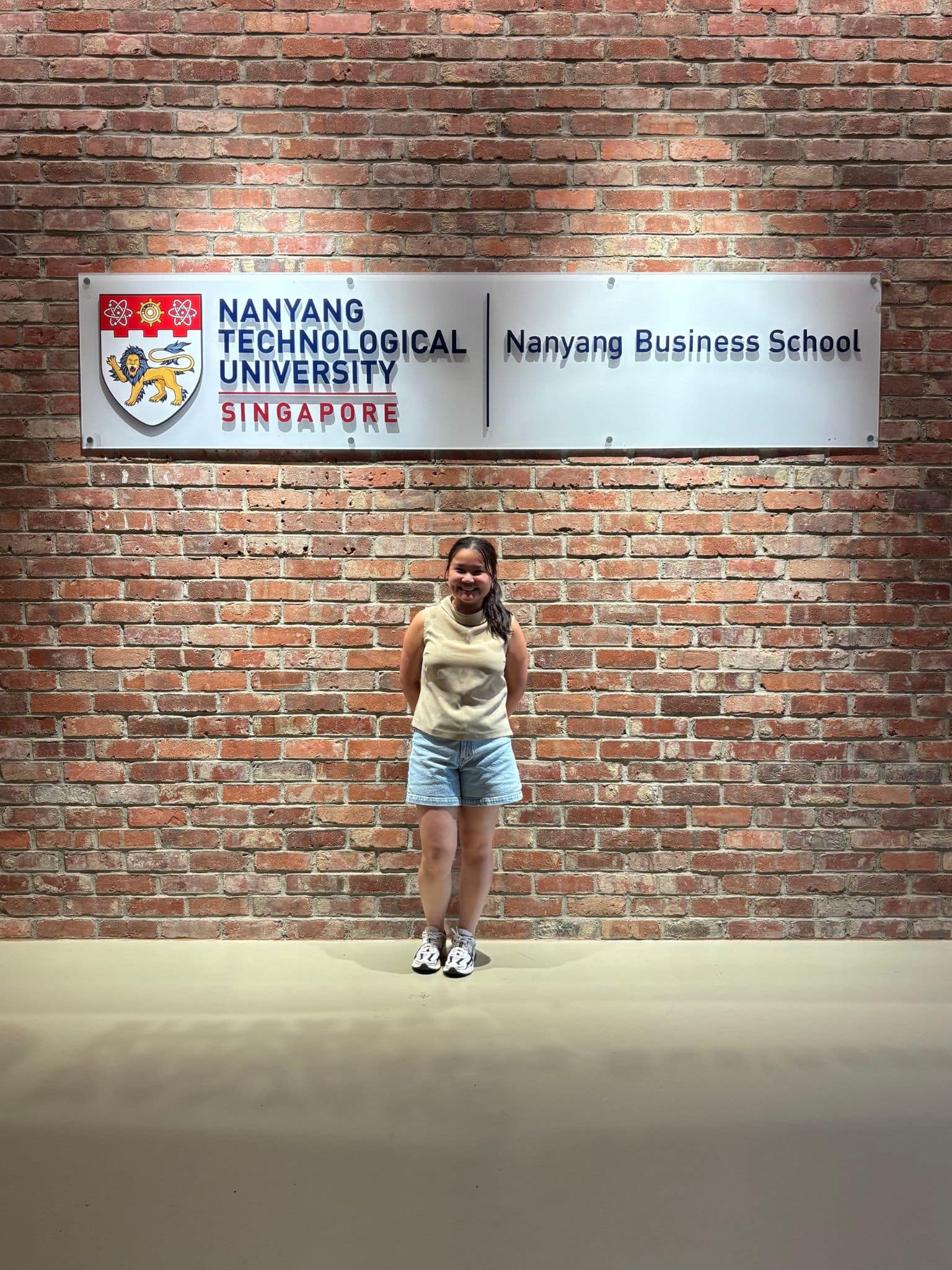 A person stands and smiles in front of a brick wall displaying a sign for Nanyang Technological University Singapore, Nanyang Business School.